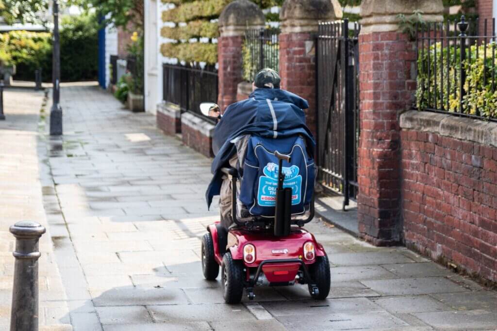 Man driving a red motility scooter on a UK street path 